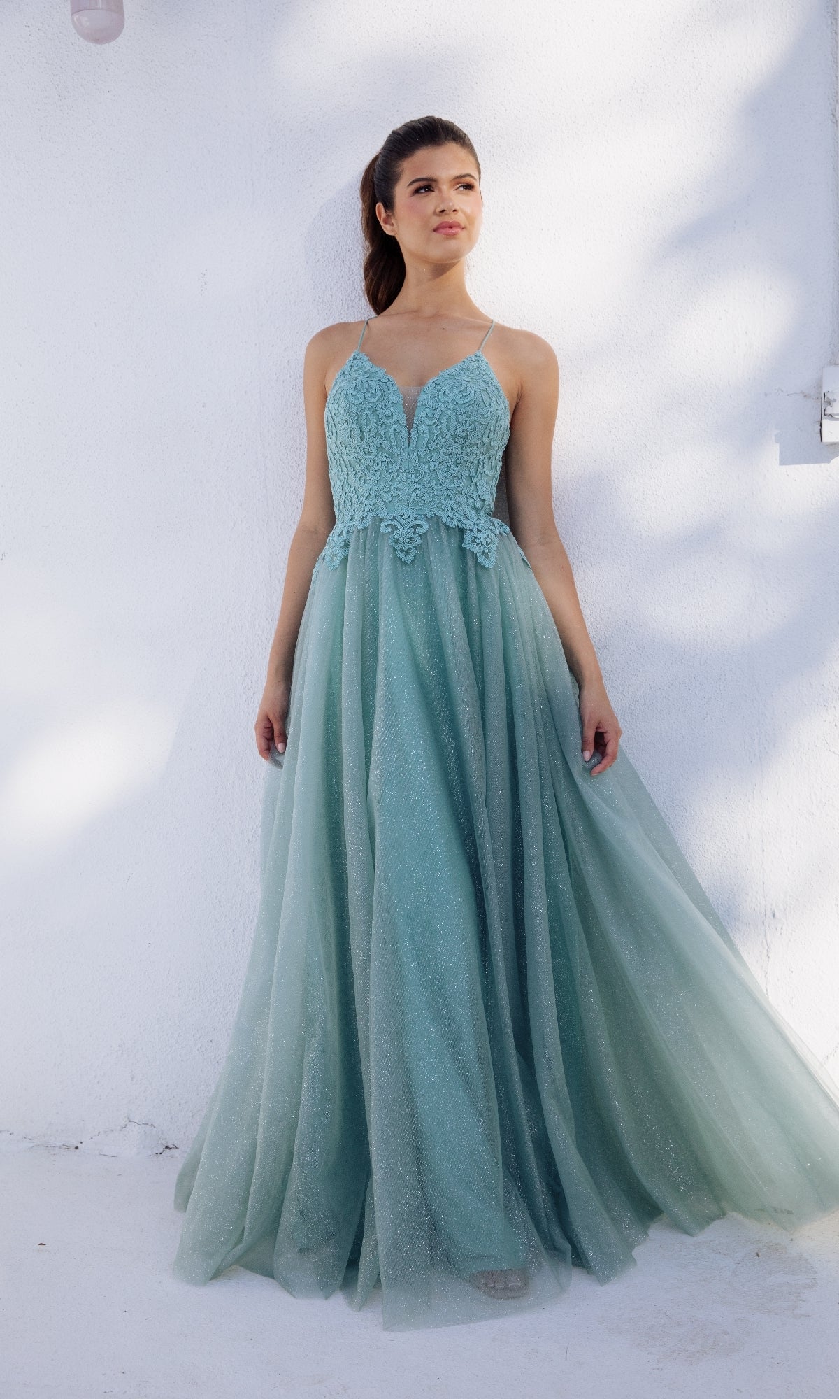A woman stands against a white wall wearing the Eureka 9901 Lace-Up Long Glitter Prom Ball Gown—a sleeveless light blue dress featuring a fitted lace bodice and a flowing tulle skirt.